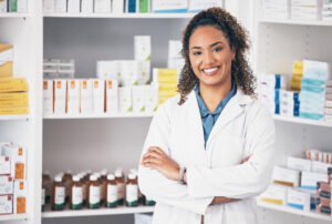 Pharmacy, pharmacist or portrait of woman with arms crossed or smile in customer services or clinic. Healthcare help desk, wellness or happy doctor smiling by medication on shelf in drugstore